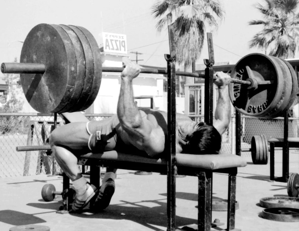 Franco Columbu performing a shirtless outdoor barbell bench press