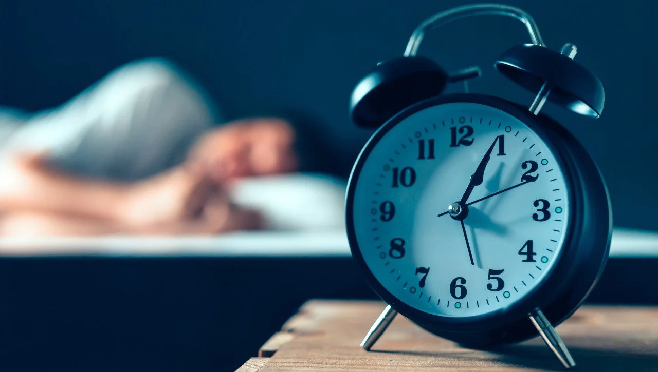 Clock on nightstand with person sleeping in the background