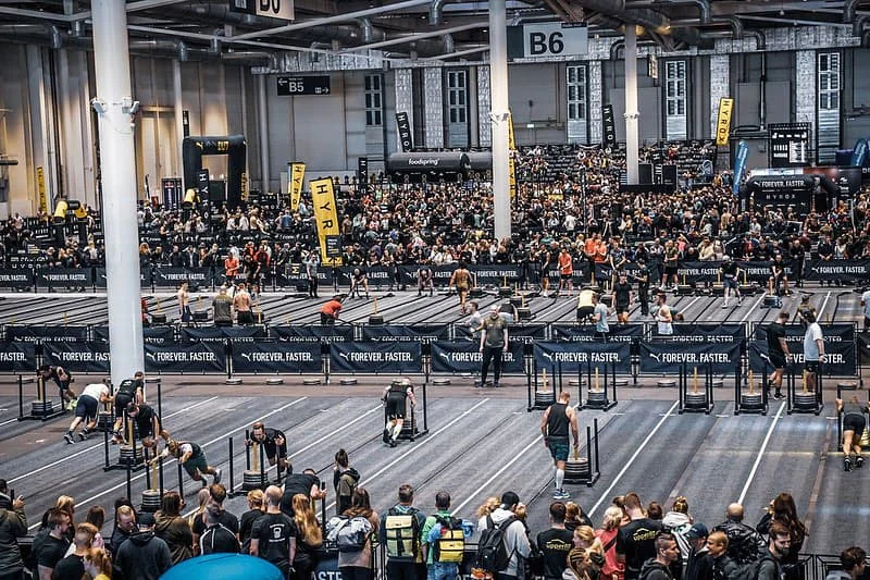 Overhead view of a Hyrox racecourse inside a large indoor arena, displaying the 1km running track and all eight workout stations, including SkiErg, Sled Push, Sled Pull, Burpee Broad Jumps, Rowing, Farmers Carry, Sandbag Lunges, and Wall Balls. Directional arrows and labels highlight the race flow.